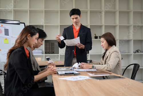 Frustrated Asian Business Manager Standing at Meeting Table Crumpling Paper While Colleagues Sit Stressed in Office, conveying workplace stress and conflict in a modern office setting
