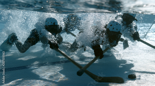 underwater hockey players in pool fins and masks scrambling for puck along tiled floor, bubbles rising around helmeted heads, surface ripples distorting overhead spectator view,