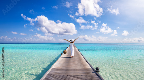 anoramic view of a happy woman in a white summer dress walking over a long pier over turquoise ocean in the Maldives islands as a travel concept