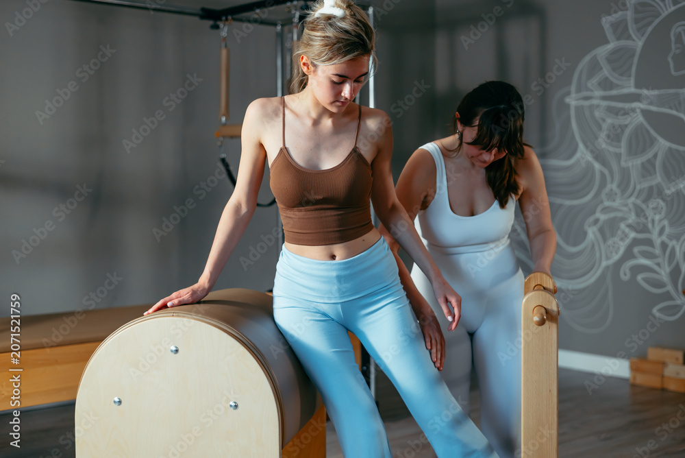 Naklejka premium Two women engage in a pilates session in a fitness studio, with one woman assisting the other on a reformer machine