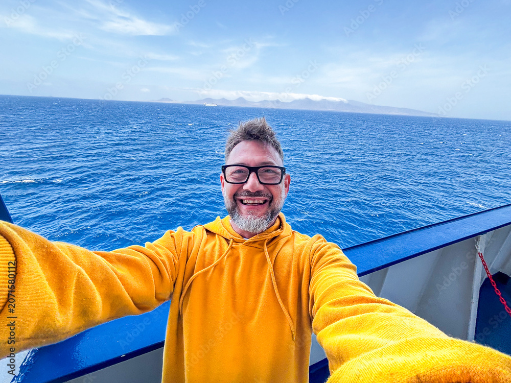 Fototapeta premium Smiling man in yellow hoodie takes a selfie on a boat with deep blue ocean and distant islands, conveying travel, happiness and adventure on a sunny day at sea