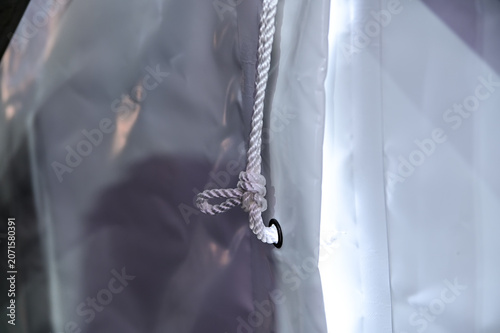 Fotografie close-up photograph focusing on a white braided nylon rope tied into a knot through a black metal grommet on a translucent plastic or vinyl sheet