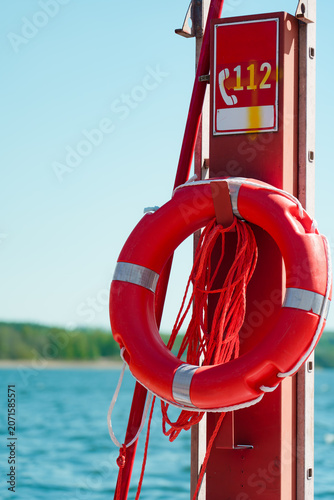 Obraz na plátne Bright red lifebuoy hangs on marina post with 112 emergency sign, rope, and meta