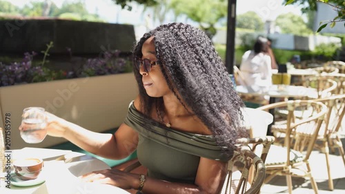 Young and beautiful black woman drinking water in a cafe and reading a book on a sunny and windy day