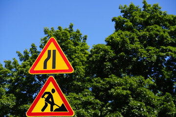 Road warning signs with lane narrowing and construction symbol against green trees and blue sky