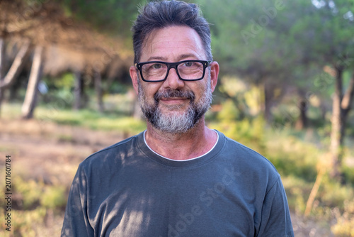 Middle-aged man with glasses and beard smiling outdoors in a casual t-shirt, relaxed portrait in a sunlit forest setting conveying warmth, confidence and friendly approachable personality.