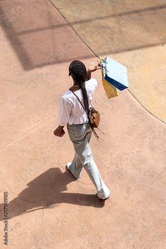 top view of afro woman with long dark braids, wearing a white lace blouse and jeans, standing with bags on a street or shopping center