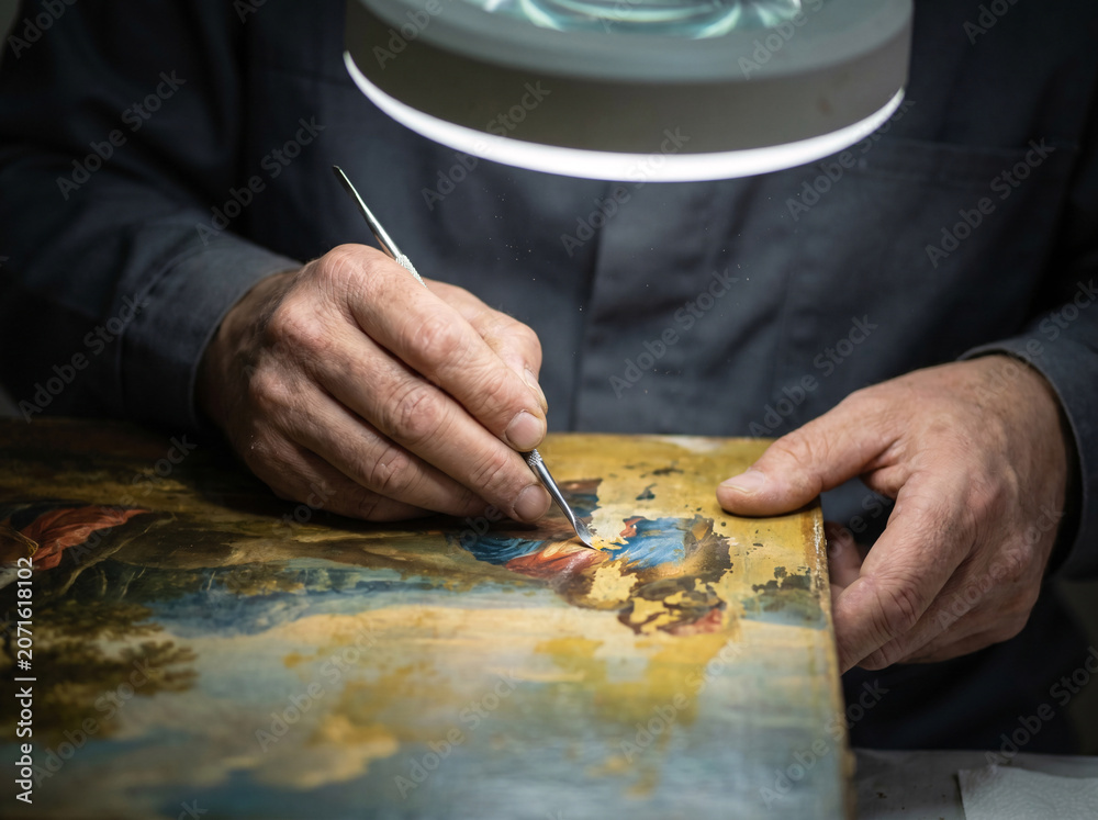Fototapeta premium Art restorer's hands working on darkened old painting as original vivid colors emerge and brighten beneath specialized tool revealing centuries hidden beauty