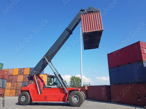 Quadro su tela Reach stacker lifting a shipping container at a busy cargo terminal, illustrating global logistics, container handling, freight transportation, port operations, and supply chain efficiency