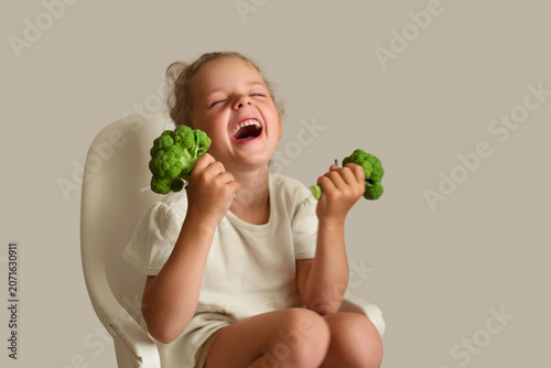 A young girl sits on a chair, holding two pieces of broccoli in her hands. She is laughing and shows joy while surrounded by a simple indoor setting