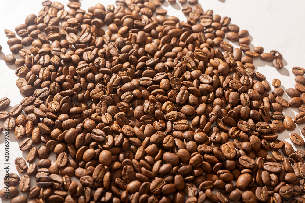 Fototapeta premium A pile of roasted coffee beans sits on a white background forming a natural mound shape. The heap of dark beans spills slightly against a clean light surface. Concept of coffee