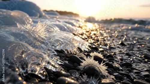 Gentle waves flow rhythmically over smooth pebbles as the sun sets on the horizon, casting golden light across the shimmering surface of the water, creating a tranquil scene filled with beauty.