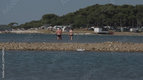 Summer scene in Medulin, Croatia, showing two people wading through shallow Adriatic waters near a rocky shoreline. A seaside campsite and pine forest form the backdrop, capturing a relaxed holiday