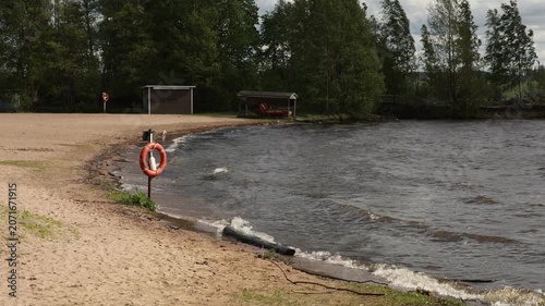 A windy day at a beach in Tampere, Finland, showing choppy water with waves hitting the sandy shore. A lifebuoy stands on the beach, with some wooden buildings and trees in the background. The sky is