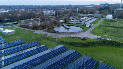 Aerial view of a wastewater treatment plant with circular clarifier tanks, water basins, technical infrastructure and large solar panel fields. Industrial water management facility.