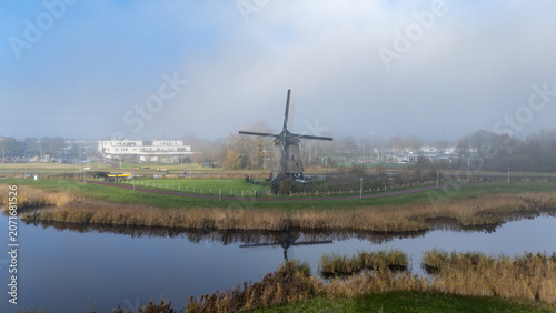 Scenic view of a traditional Dutch windmill beside calm water with reeds, grass and soft mist in the background. Peaceful countryside landscape with natural reflection and atmospheric morning light.