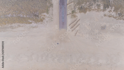 Top down aerial view of a sandy beach with a wooden boardwalk leading through coastal dunes. Minimal seaside landscape with soft sand textures, dune grass and natural patterns seen from above.