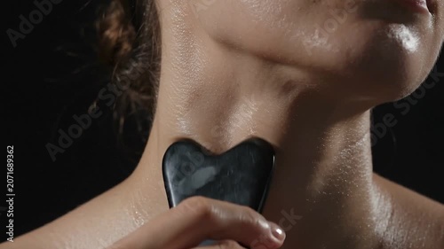 A close up look of a woman's neck and jawline while practicing a skincare massage using a black heart shaped gua sha stone tool