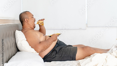 Lazy Overweight Asian Man Eating Hamburger in Bed, Unhealthy Lifestyle Concept