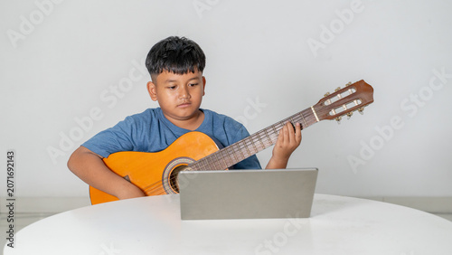 Focused Young Asian Boy Learning to Play Acoustic Guitar with Laptop Online Lesson
