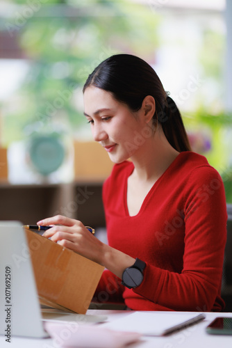 Young Asian woman preparing a parcel for shipping, labeling a brown cardboard box with a pen, managing her small business, and handling e-commerce operations in a bright home office