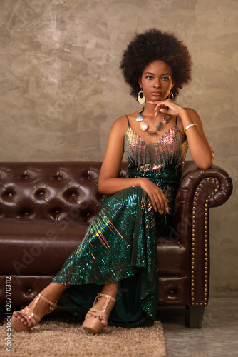 Portrait of an elegant African woman with an Afro hairstyle, wearing a long dress, sitting on a vintage-style sofa. Vertical image.