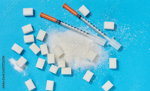 Two insulin syringes and scattered white sugar on a blue background, representing the problem of diabetes and glucose dependence.