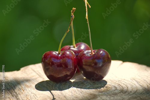 Three ripe dark cherries with stems rest on a weathered wooden surface outdoors
