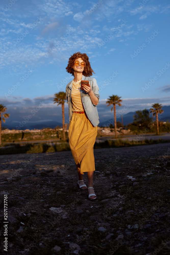Fototapeta premium A young woman with curly hair walks along a rocky beach at golden hour, wearing a mustard dress and light jacket, smiling softly as palm trees frame the scene.