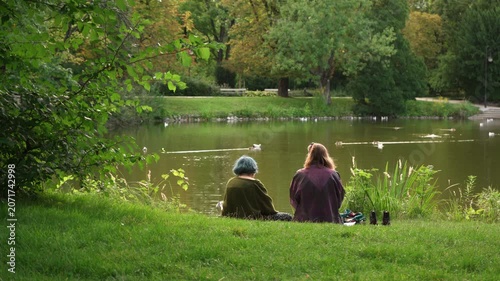 Two young lesbian women sit on grass in public park by pond enjoying outdoor leisure. Female couple or diverse friends spend weekend together reflecting closeness, intimacy and lgbt lifestyle.