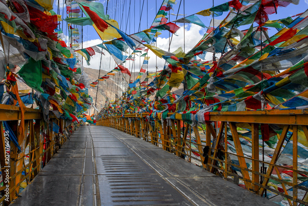Fototapeta premium Colorful Tibetan prayer flags covering a suspension bridge in a mountainous Himalayan landscape. Cultural travel destination featuring Buddhist traditions and scenic architecture.