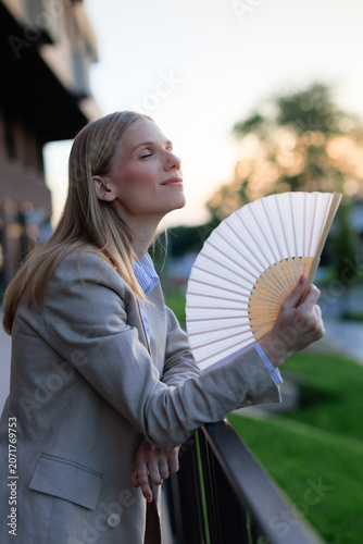 Young businesswoman cools down with a fan during heatwave while standing on balcony in evening light