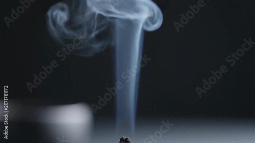 A lit incense stick smolders in a serene setting. Thin wisps of blue-grey smoke rise steadily, curling into elegant, ethereal patterns against a minimalist black background.