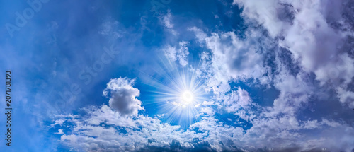 Sun with cu,ulus clouds drift and swirl across a clear, bright blue sky, creating a dynamic airy atmospheric. Shine with radial rays on center composition rainy small cloud on front of the scene.