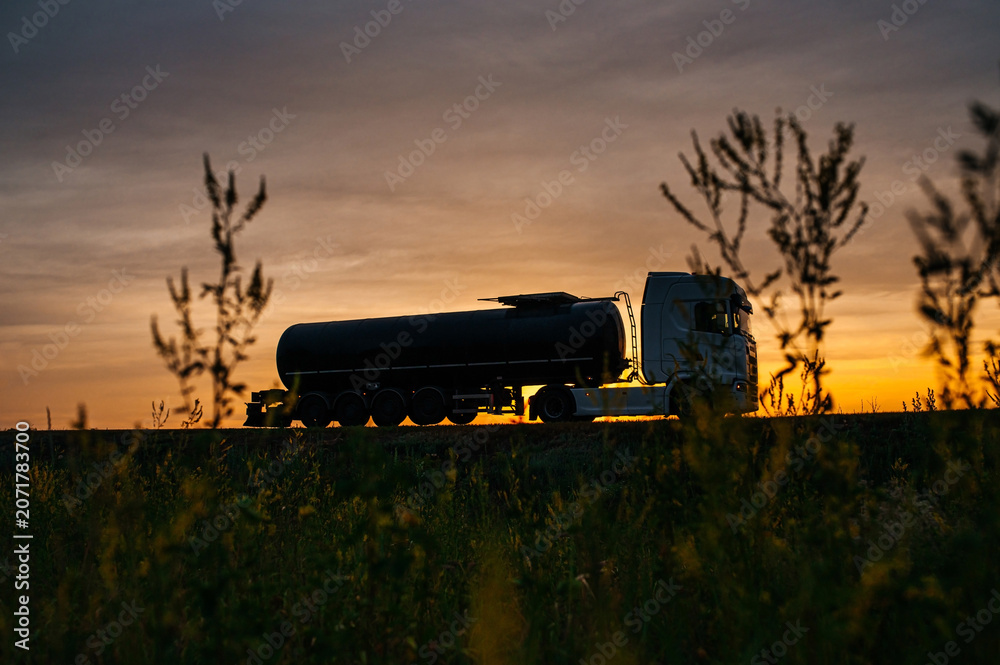 Fototapeta premium Silhouette of a fuel tanker driving along a highway at sunset in summer