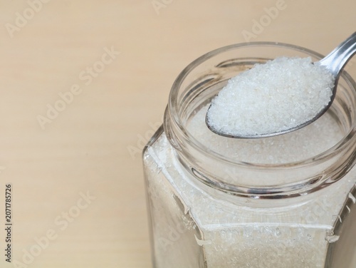Fotografie Close-up shot of white refined granulated sugar with a stainless steel spoon from a transparent glass storage jar