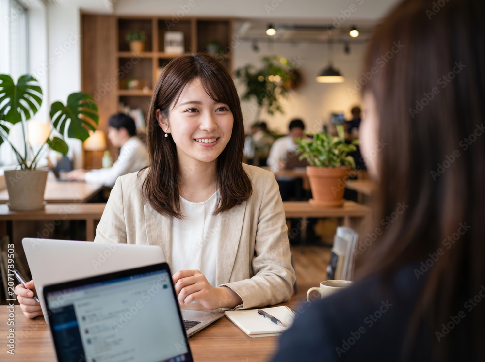 Fototapeta premium カフェスペースでPCを使い同僚と談笑するジャケットの女性