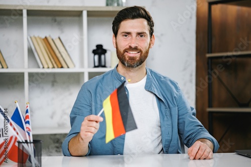 Bearded male tutor sitting at table holding flag of Germany looking at camera