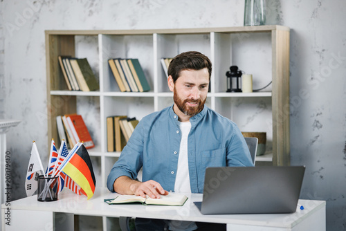 Bearded male tutor sitting at table with different flags and looking at laptop