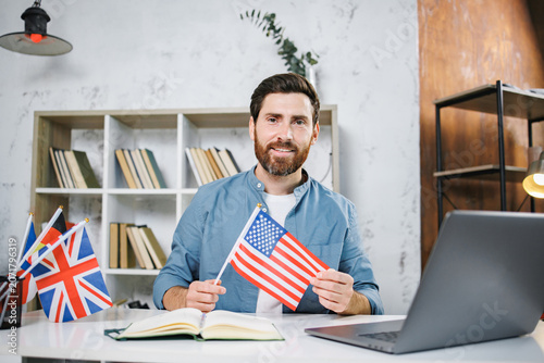 Bearded male tutor sitting at table holding flag of USA, looking at camera