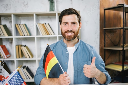 Bearded male tutor sitting at table holding flag of Germany looking at camera