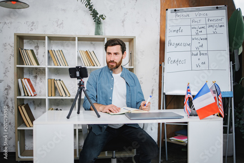 Bearded male tutor sitting at table next to whiteboard and recording a video