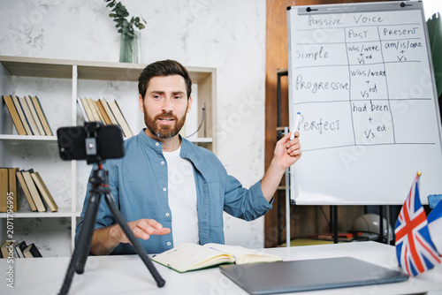 Bearded male tutor sitting at table next to whiteboard and recording a video