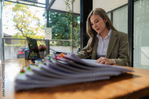 Asian businesswoman writing and checking information on paperwork with a large stack of documents on office desk.