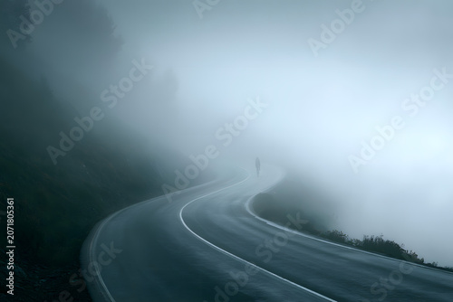 A lone cyclist on a winding mountain road enveloped in dense fog, creating a mysterious and atmospheric, serene, and solitary scene.