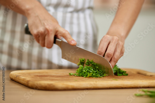Hands chopping fresh parsley or cilantro with a sharp chef knife on a wooden cutting board, preparing ingredients for cooking