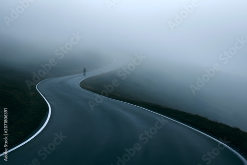 A lone cyclist on a winding mountain road enveloped in dense fog, creating a mysterious and atmospheric, serene, and solitary scene.