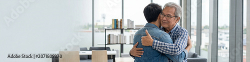 A senior man warmly embraces a younger man in a bright modern office filled with bookshelves and soft natural window light