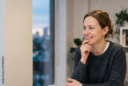 A cheerful adult woman in a warm grey sweater sits next to a window at sunset while smiling and looking away in her cozy home