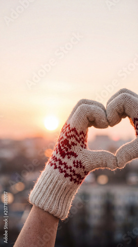 Close up of hands in cozy winter gloves forming a heart shape against a beautiful city skyline background during sunset